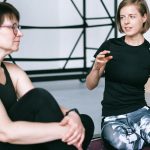 Two women in sportswear sitting on a yoga mat, having a detailed discussion in a gym.